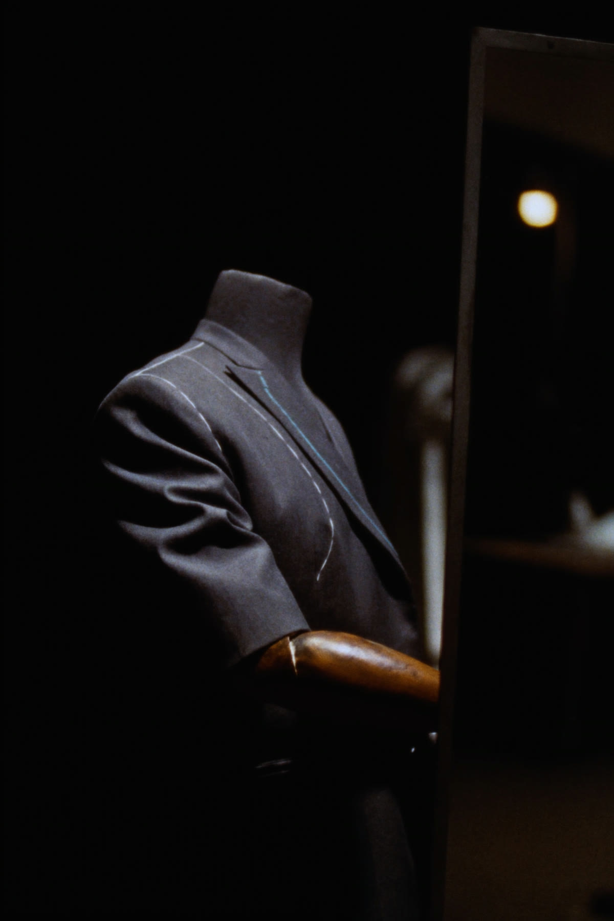 A half-finished charcoal jacket on a wooden tailor's form, basted with white thread, in front of a tall mirror.