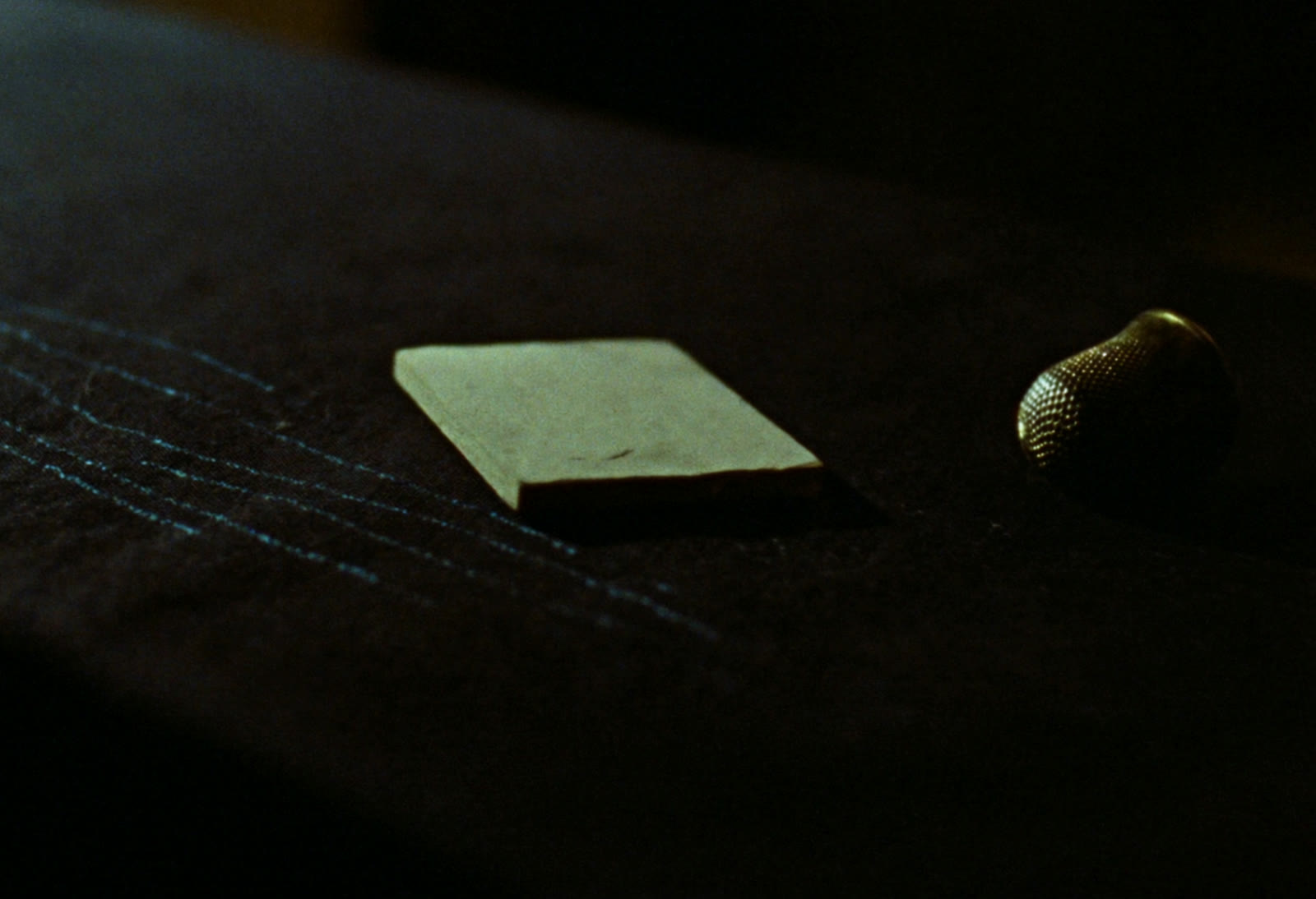 A flat piece of tailor's chalk and a brass thimble resting on charcoal wool, with faint chalk lines drawn across the cloth.