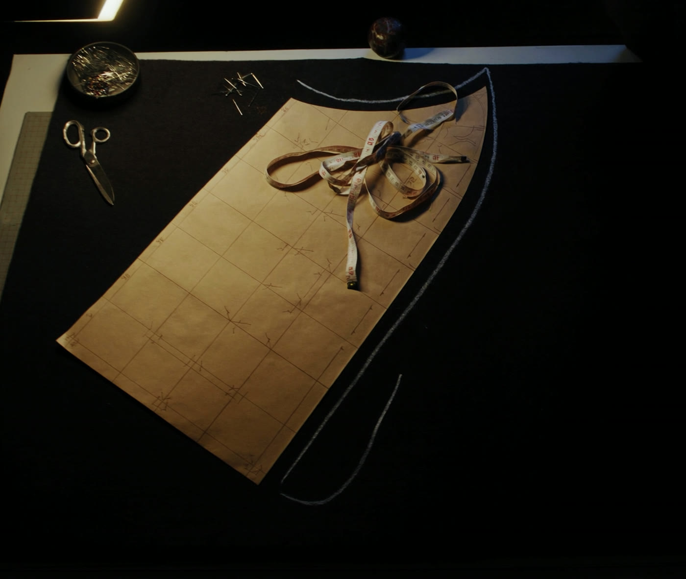 A cutting table seen from above: pattern paper with drafting lines, a coiled tape measure, shears and pins on dark cloth.